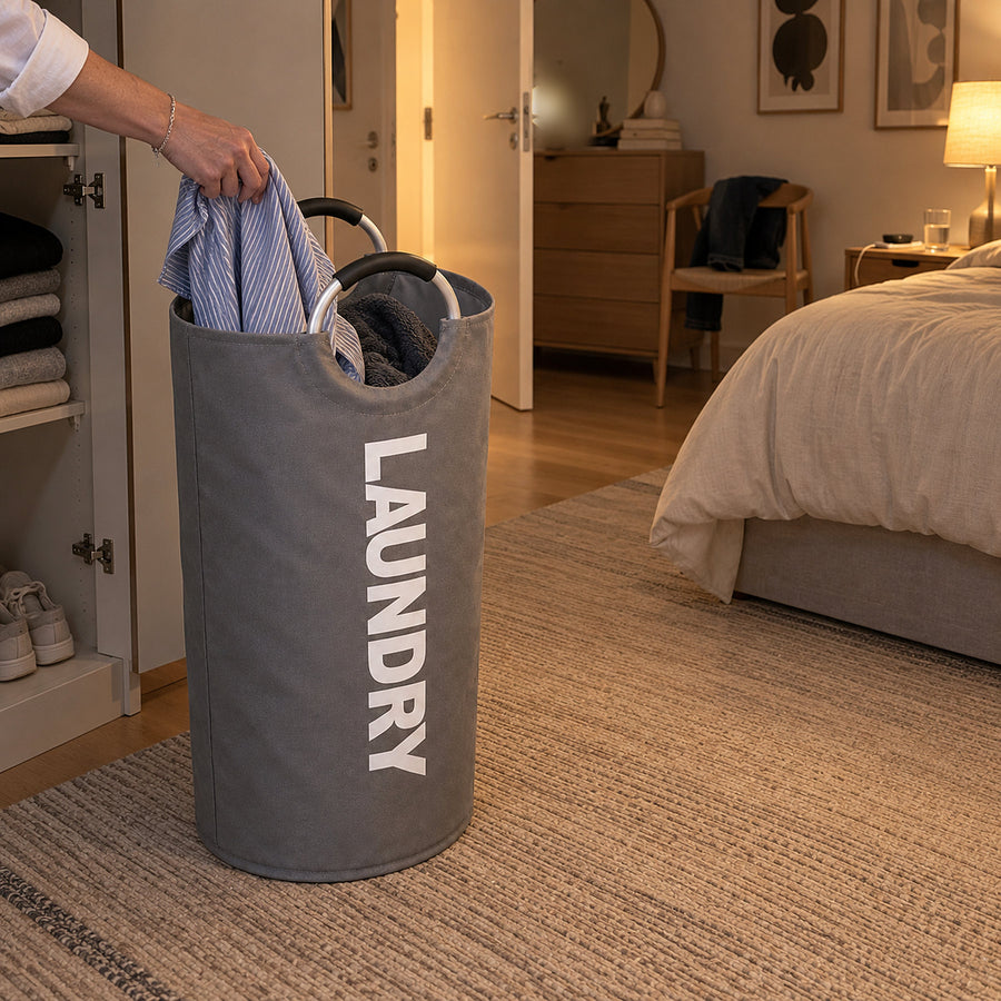 Grå hopfällbar tvättkorg med bärhandtag i ett inrett sovrum där en hand lägger en randig skjorta i korgen