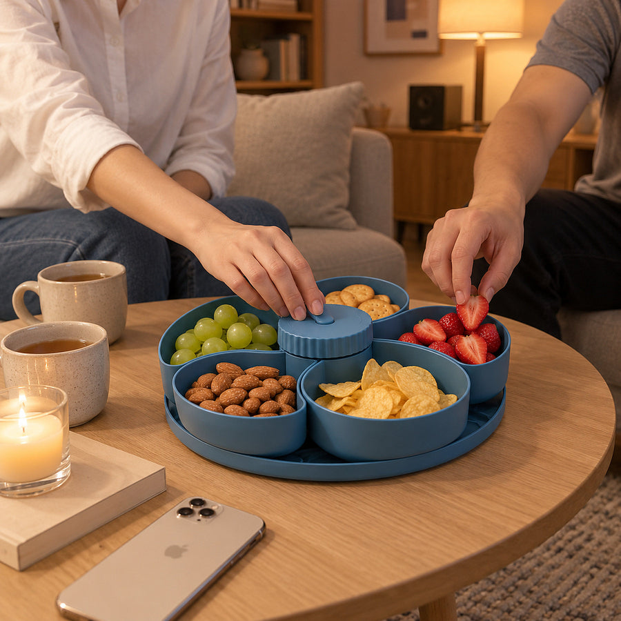 Roterande snacksbricka i blått med fem fack fyllda med vindruvor, mandel, chips, jordgubbar och kex på ett soffbord, två personer sitter i soffan