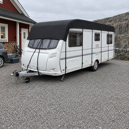 Husvagn med svart taköverdrag parkerad på en grusplan framför ett rött trähus