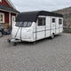 Husvagn med svart taköverdrag parkerad på en grusplan framför ett rött trähus