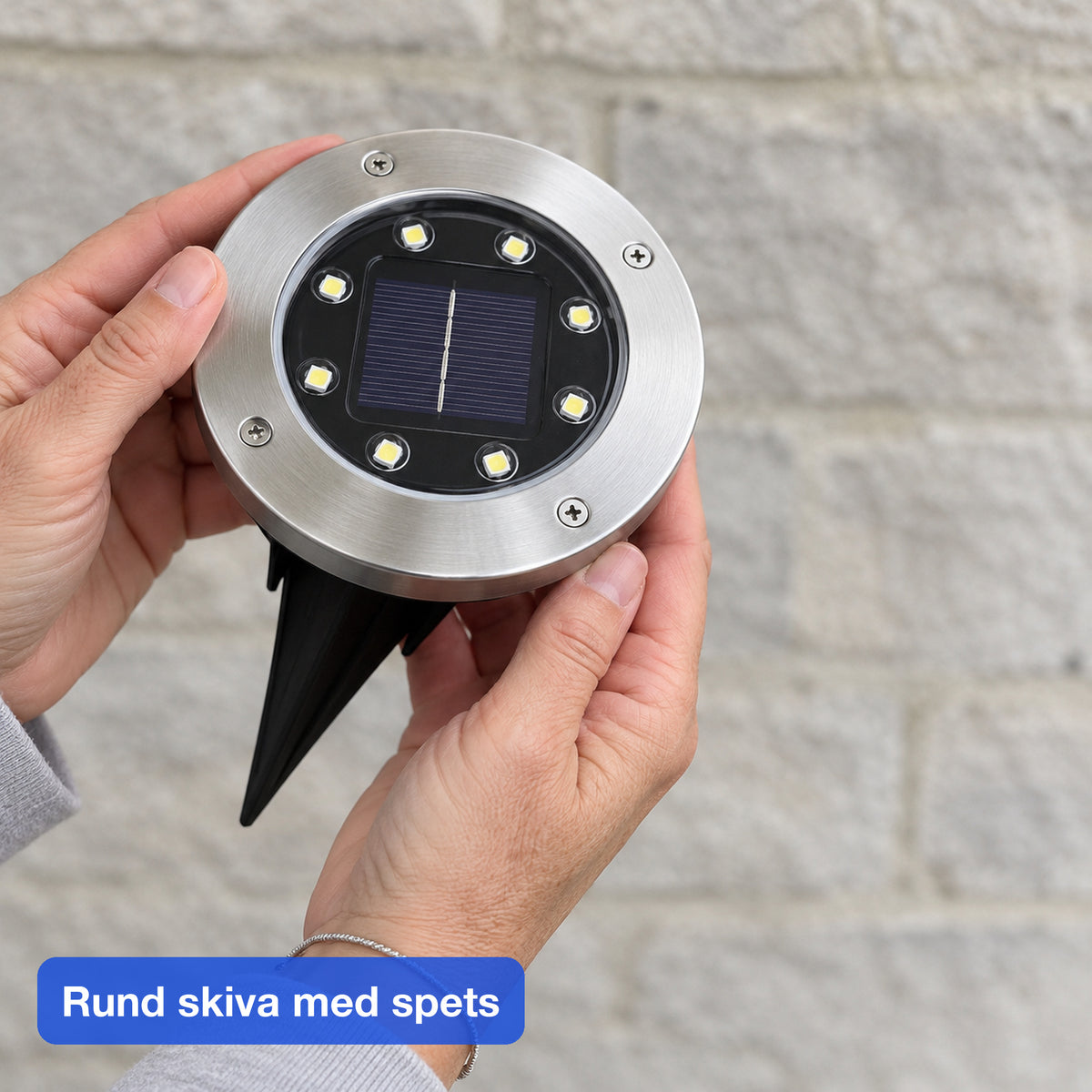 Två händer håller en rund marklampa med metallring, solpanel i mitten och svart spets