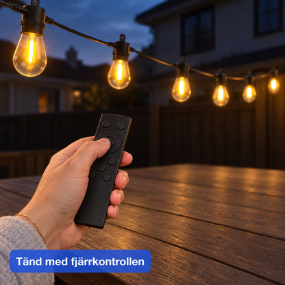 Hand riktar en svart fjärrkontroll mot en tänd ljusslinga över ett utebord i skymning