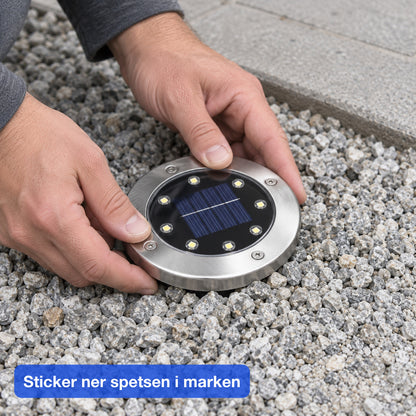 Två händer trycker ner en rund marklampa med metallring i grus