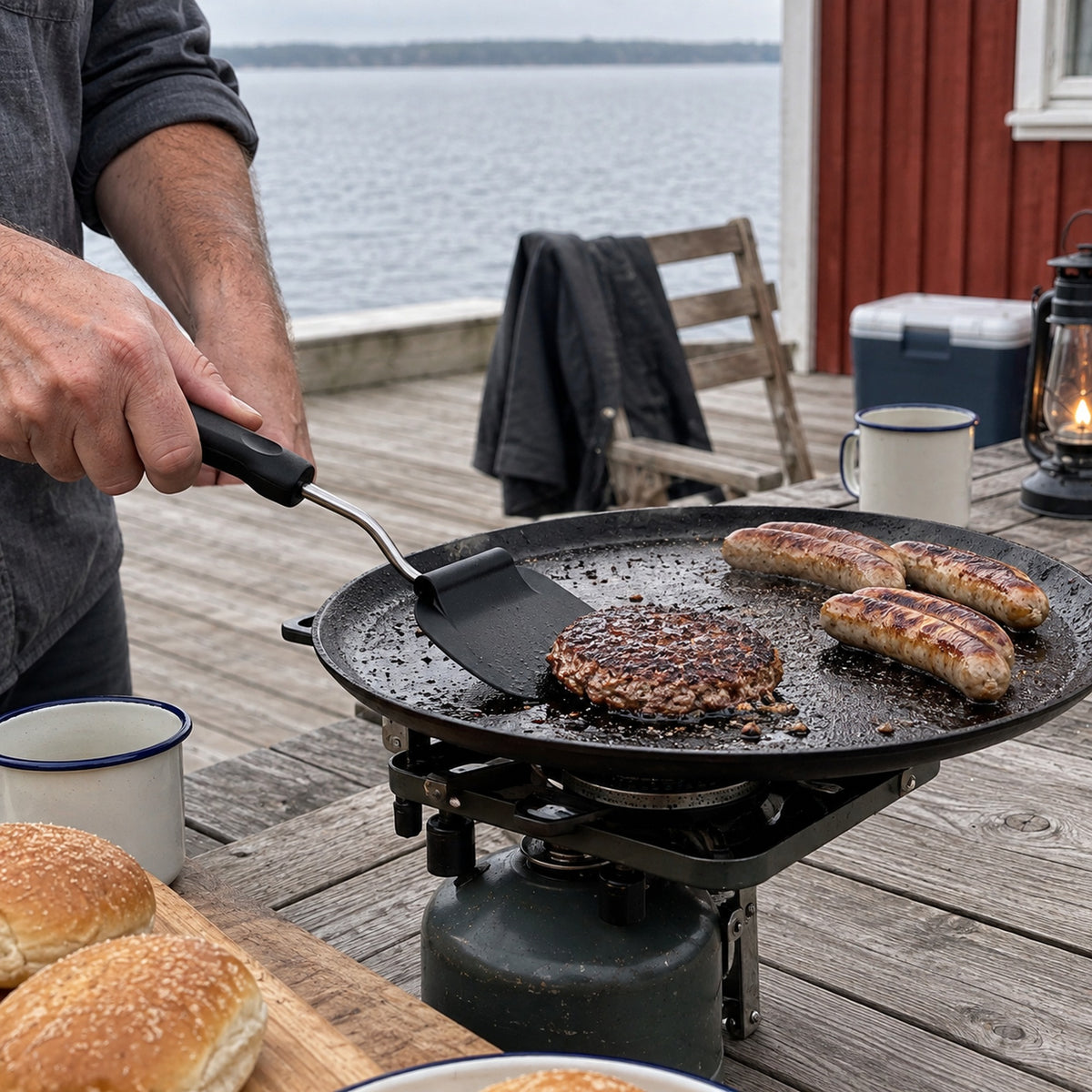Vinkelbar stekspade med svart blad som vänder en hamburgare på en grillplatta på en träaltan vid en sjö