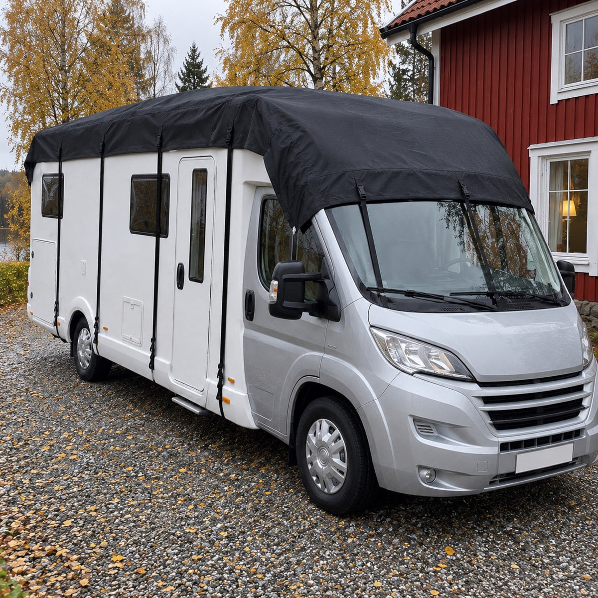 Vit husbil framifrån med svart taköverdrag och remmar på en grusplan en höstdag