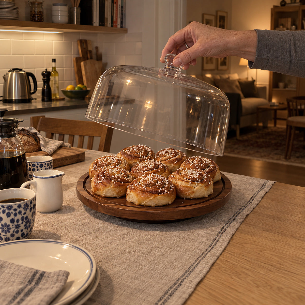 Hand lyfter den genomskinliga kupolen från träbrickan med nybakade kanelbullar