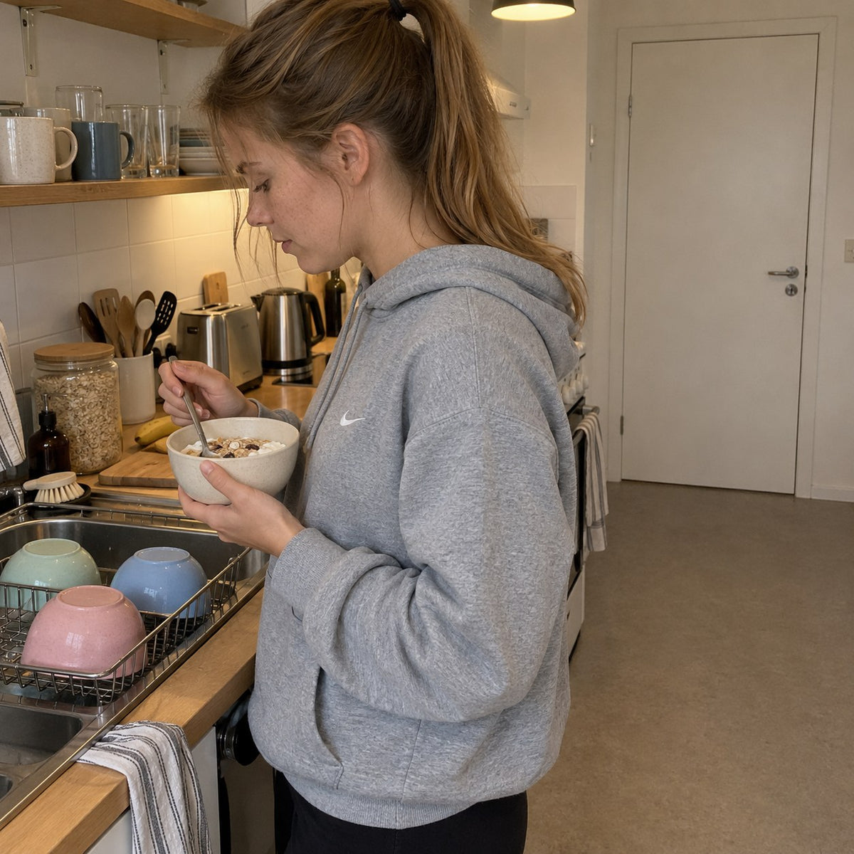 Ung kvinna äter müsli ur en skål i ett delat studentkök medan de andra skålarna står på diskstället
